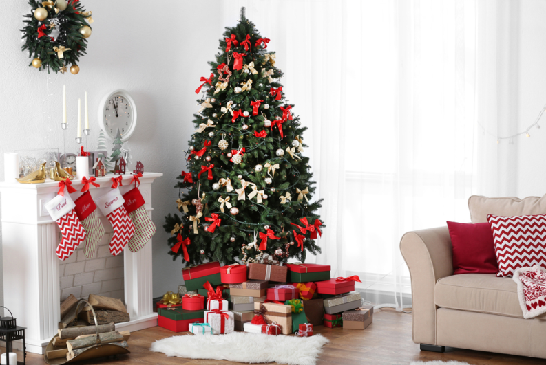 Cây thông Noel treo nhiều nơ đỏ, quả cầu, dưới là nhiều hộp quà đặt cạnh lò sưởi, một bên là ghế sofa.